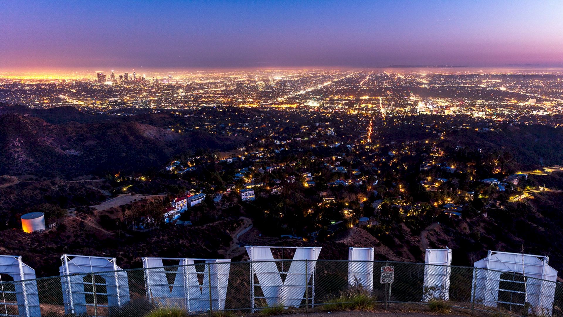 The image shows the Hollywood Sign from behind, overlooking a brilliantly lit cityscape at dusk, with the urban sprawl extending to the horizon.