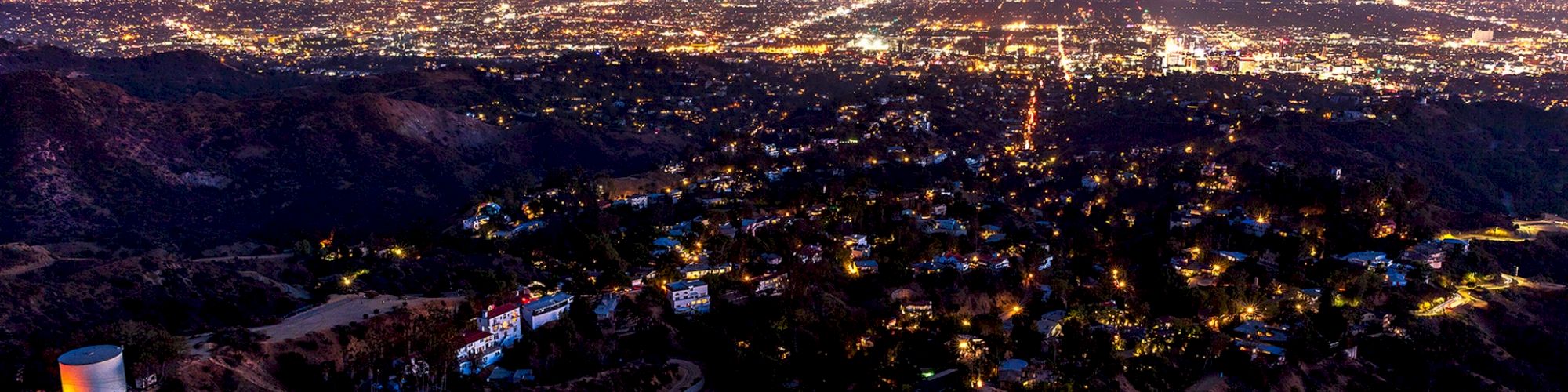 The image shows the Hollywood Sign from behind, overlooking a brilliantly lit cityscape at dusk, with the urban sprawl extending to the horizon.