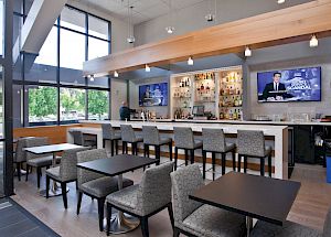 The image shows a modern bar with a TV, bar stools, and several seating areas with tables and chairs. It has large windows and a sleek design.