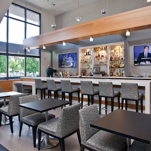 The image shows a modern bar with a TV, bar stools, and several seating areas with tables and chairs. It has large windows and a sleek design.