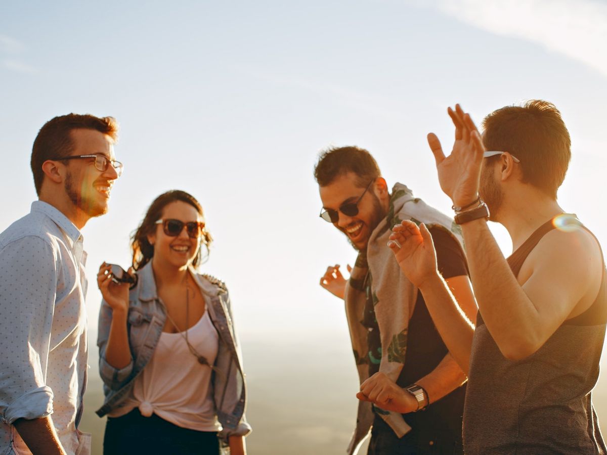 Four people are standing outdoors, laughing and enjoying the moment under the sun. They appear to be engaging in a cheerful conversation.