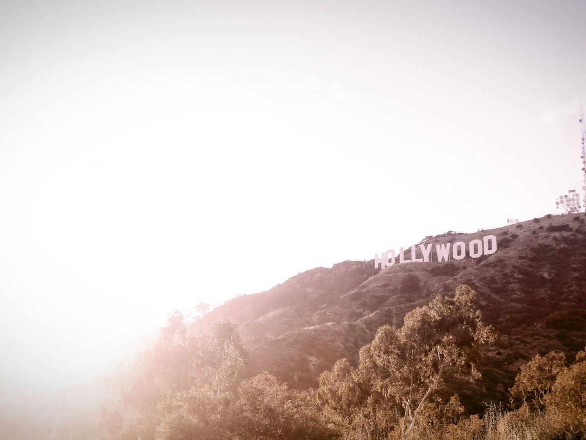 The image shows the iconic Hollywood Sign on a hillside with a communication tower nearby, surrounded by trees under a bright, hazy sky.