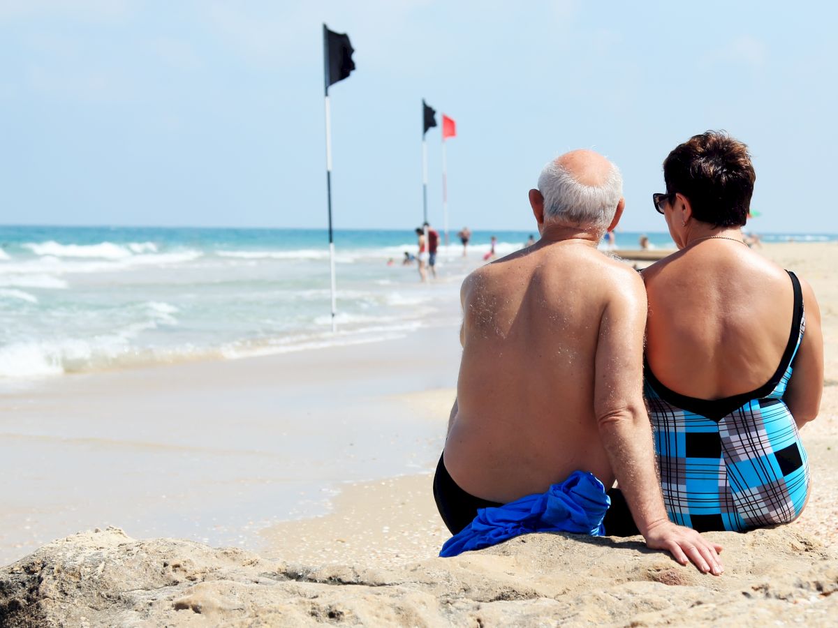 A couple sits on a beach facing the ocean with warning flags in the distance, enjoying a peaceful moment together.