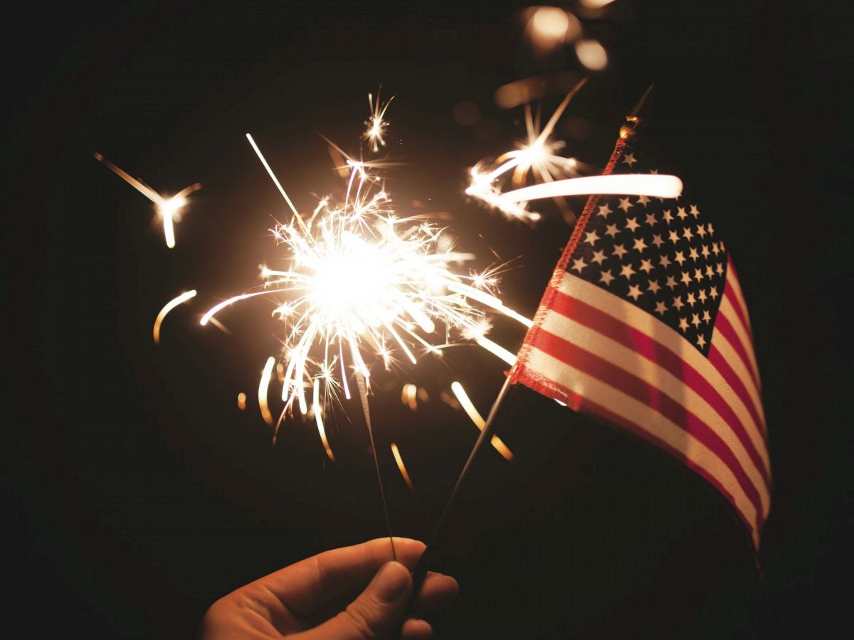 A hand holds a sparkler and a small American flag with sparks flying, set against a dark background.