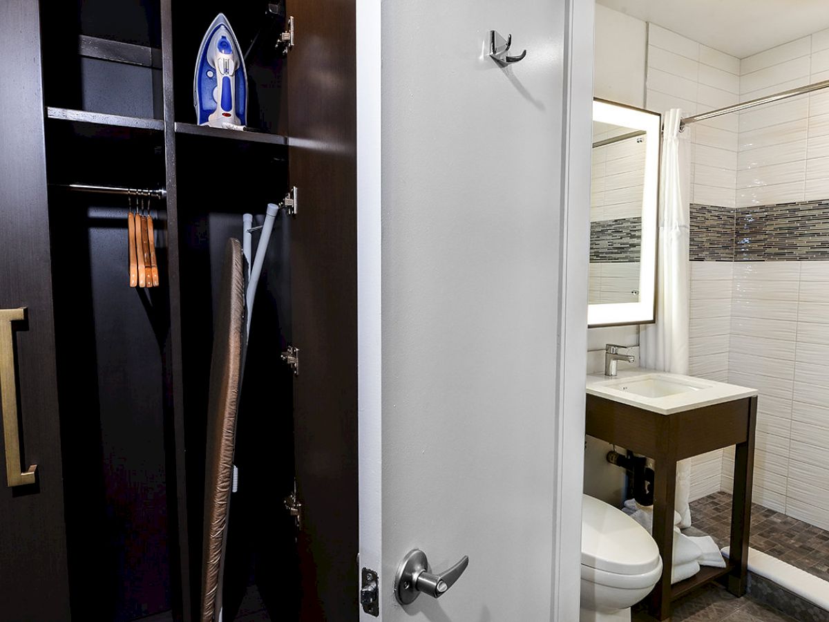 A bathroom with a sink, toilet, and mirror is visible through an open door next to a closet with an ironing board, iron, and hangers inside.