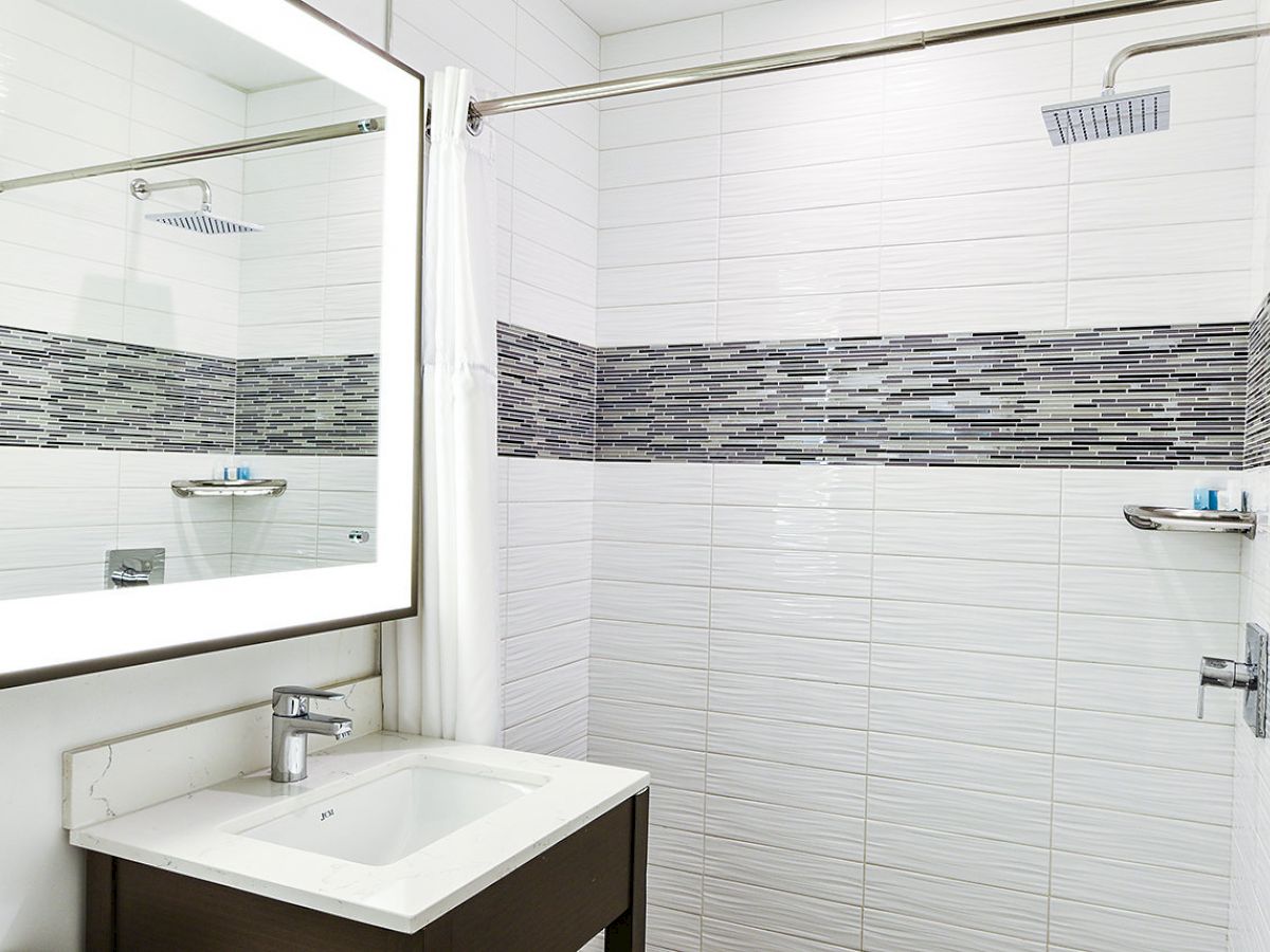 A modern bathroom with a sink, a large mirror, a shower area with a curtain, and a tiled wall with a horizontal mosaic stripe, all in white and gray.