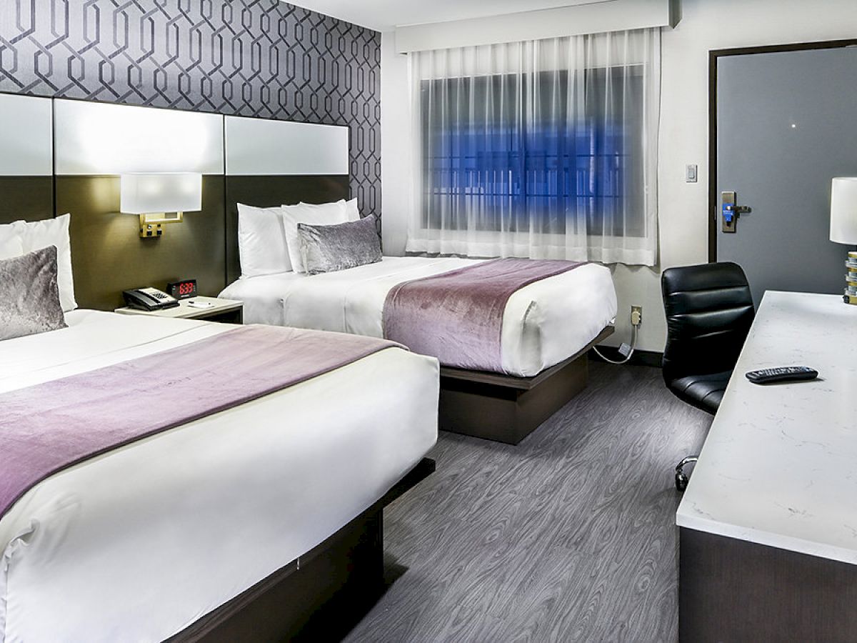 This image shows a modern hotel room with two beds, a desk with a chair, a TV remote, a lamp, and a window with curtains.