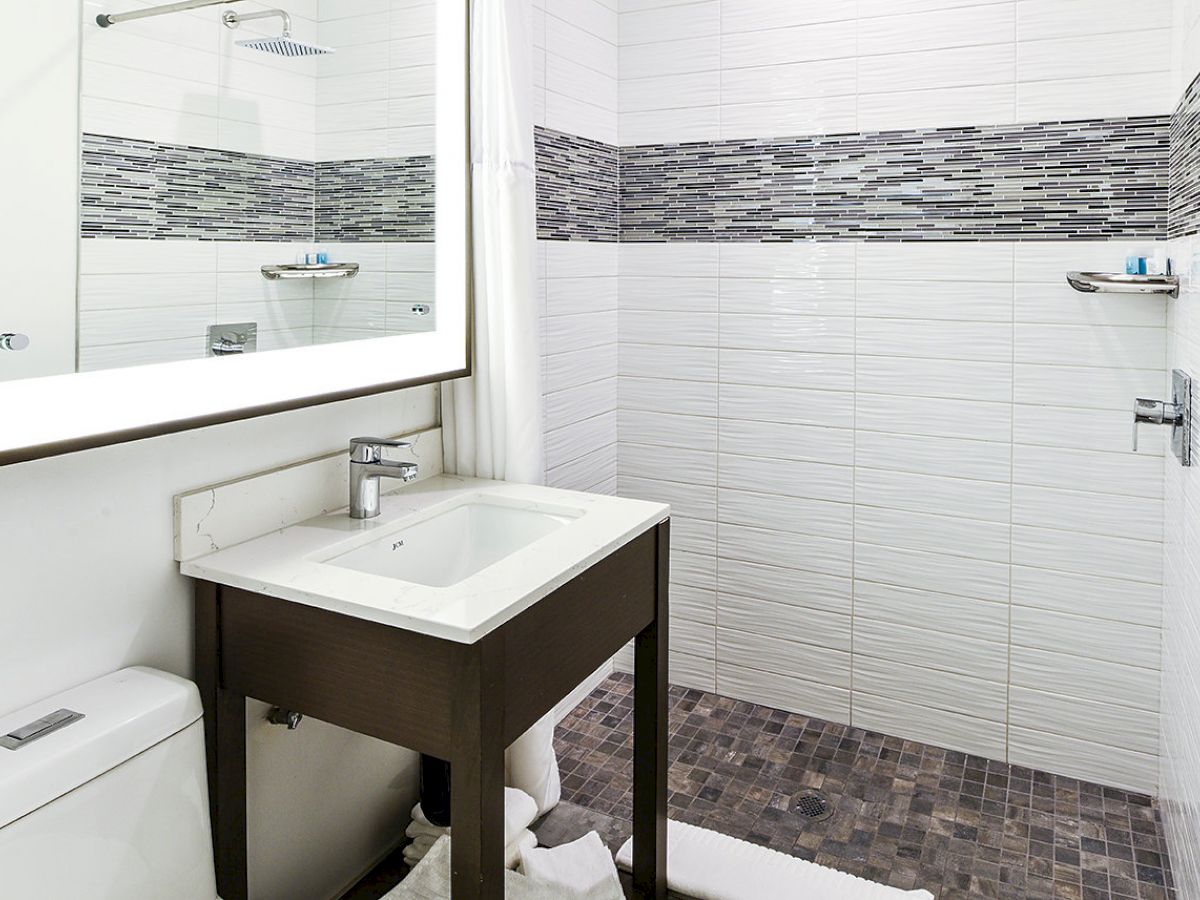 This image shows a modern bathroom with a large mirror, sink, toilet, and a walk-in shower with grey tile accents. Towels are on the floor.