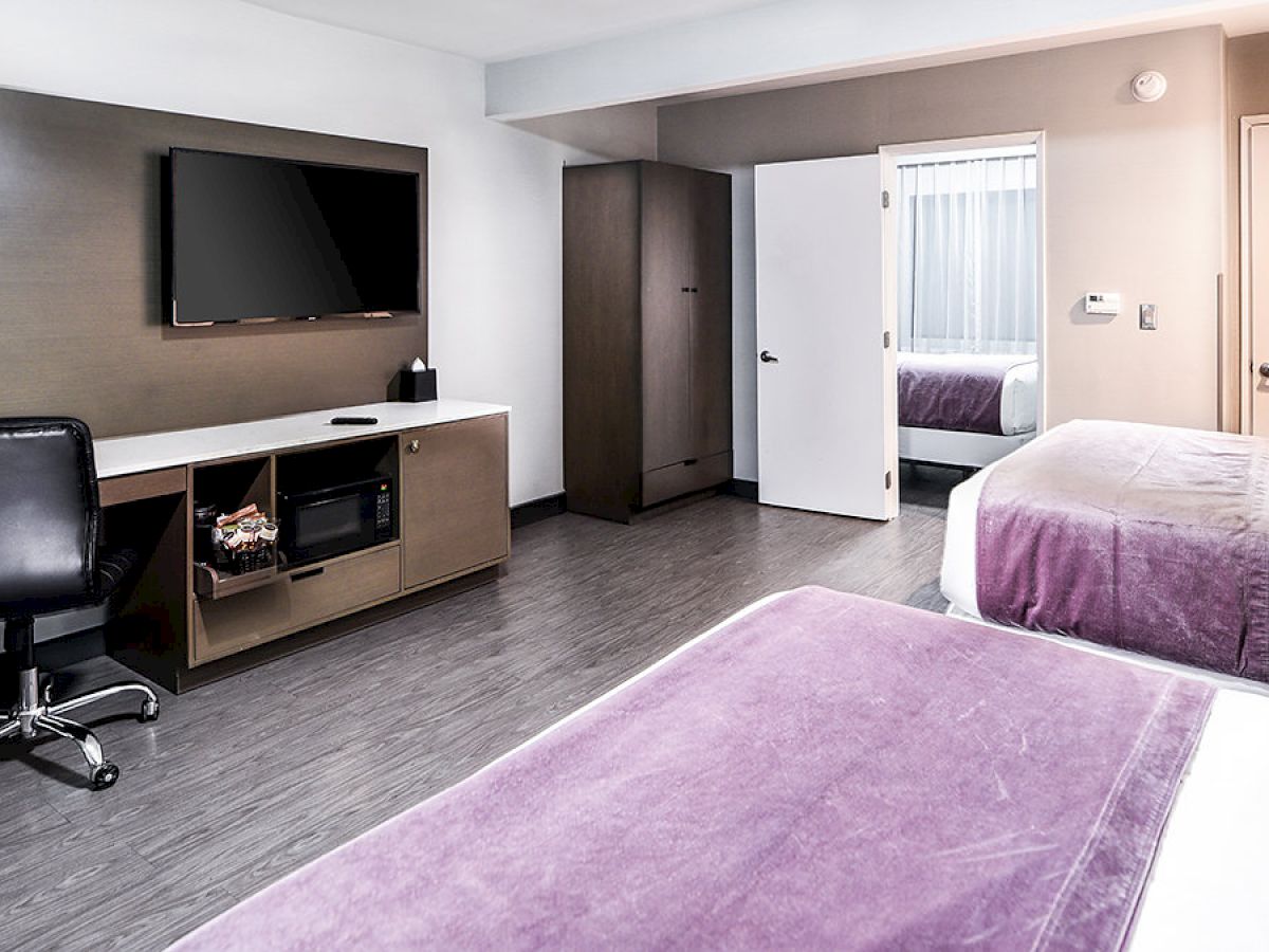 A hotel room featuring two beds with purple bedding, a desk with a chair, a wall-mounted TV, a microwave, and a fridge, in a modern decor.