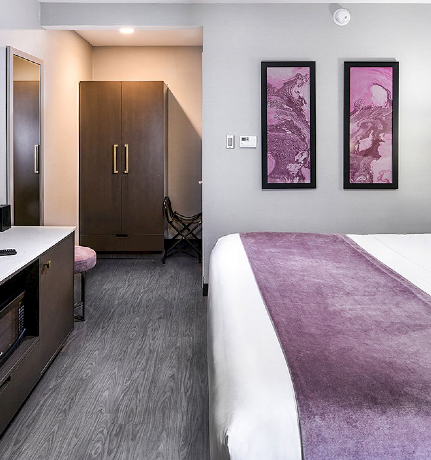 The image depicts a modern hotel room with a large bed, a purple blanket, a wall-mounted TV, a desk, and two framed artworks on the wall.