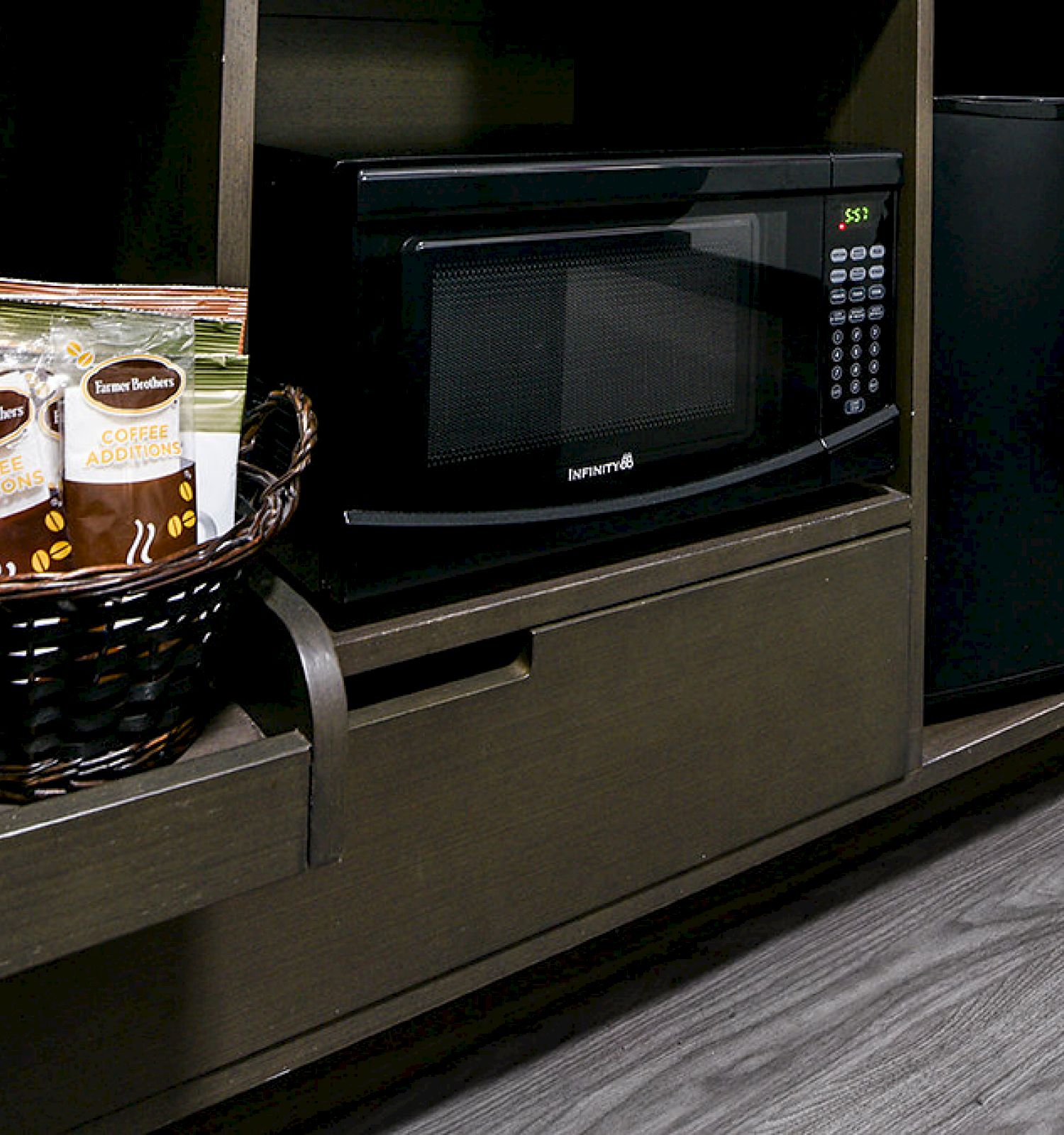 The image shows a shelf with a microwave, a small fridge, a basket with coffee supplies, and a coffee maker on a wooden floor.