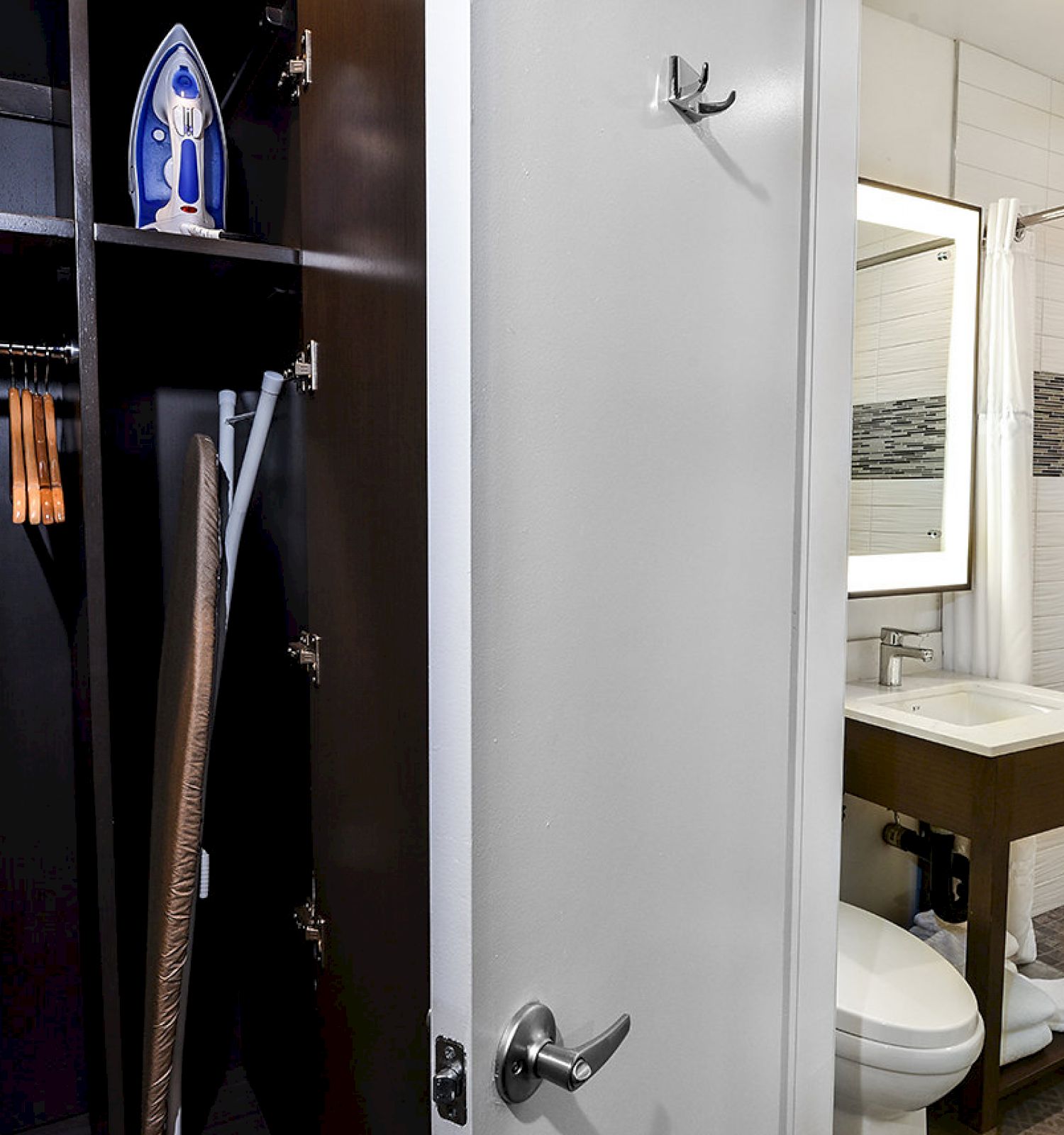 The image shows a closet with an iron, broom, and hangers, next to a bathroom with a sink, mirror, and a partially visible toilet and shower.