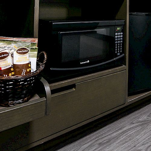 The image shows a small kitchenette area with a coffee maker, a basket with packaged coffee, a microwave oven, and a mini-fridge.