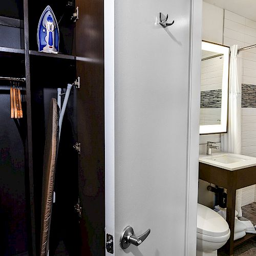 The image shows an open closet with an iron and ironing board next to a bathroom with a sink, toilet, and shower.