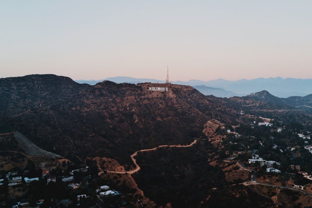 Aerial view of a mountainous area with the iconic Hollywood sign in the distance, surrounded by winding roads and scattered houses, at sunset.