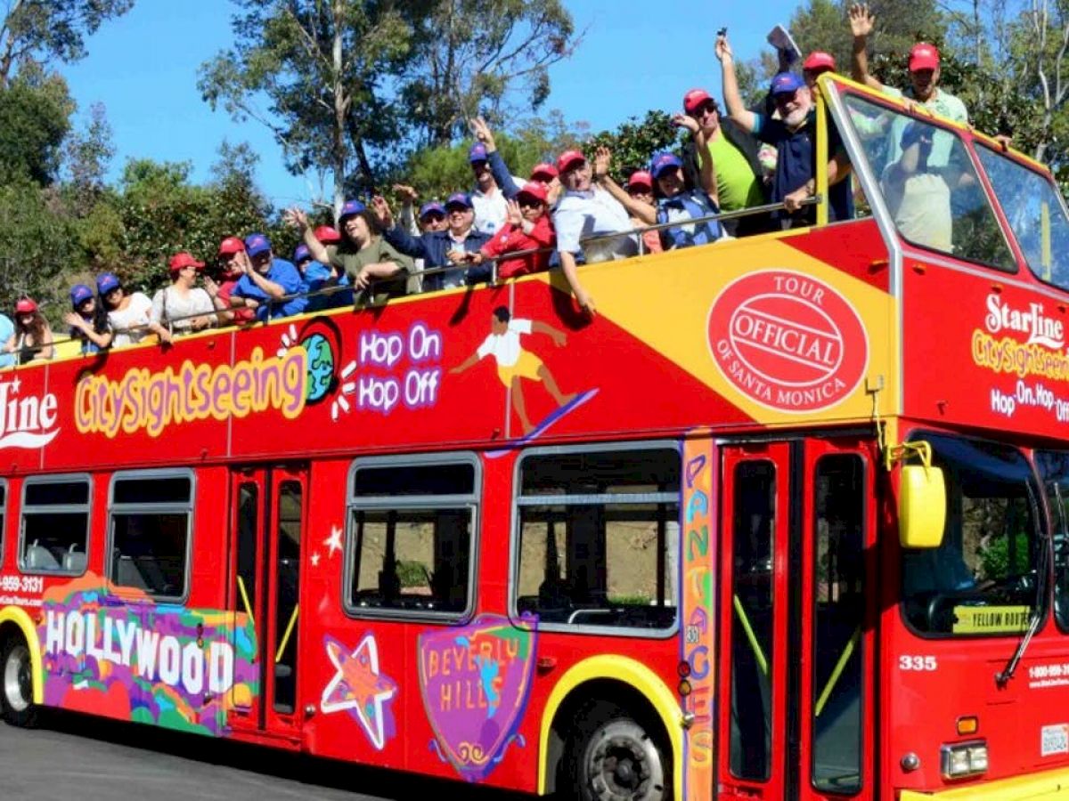 A double-decker bus filled with tourists has "City Sightseeing" written on it, and people are waving from the top deck enjoying the ride through Hollywood.