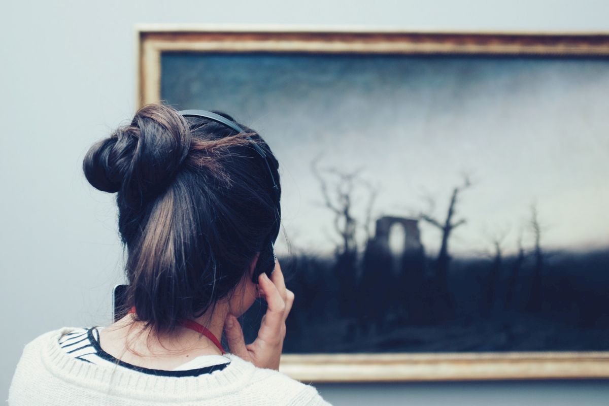 A person with a bun hairstyle, seen from behind, looking intently at a framed painting of a desolate landscape with barren trees.