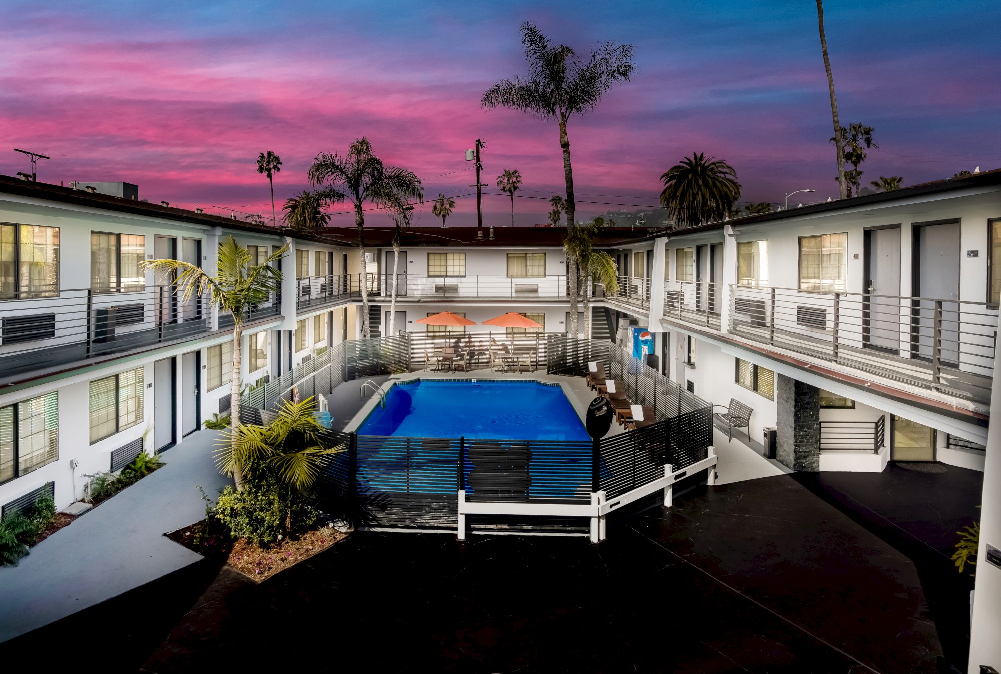 An evening view of a U-shaped building with balconies, a central pool area, and palm trees against a vibrant pink and purple sky.