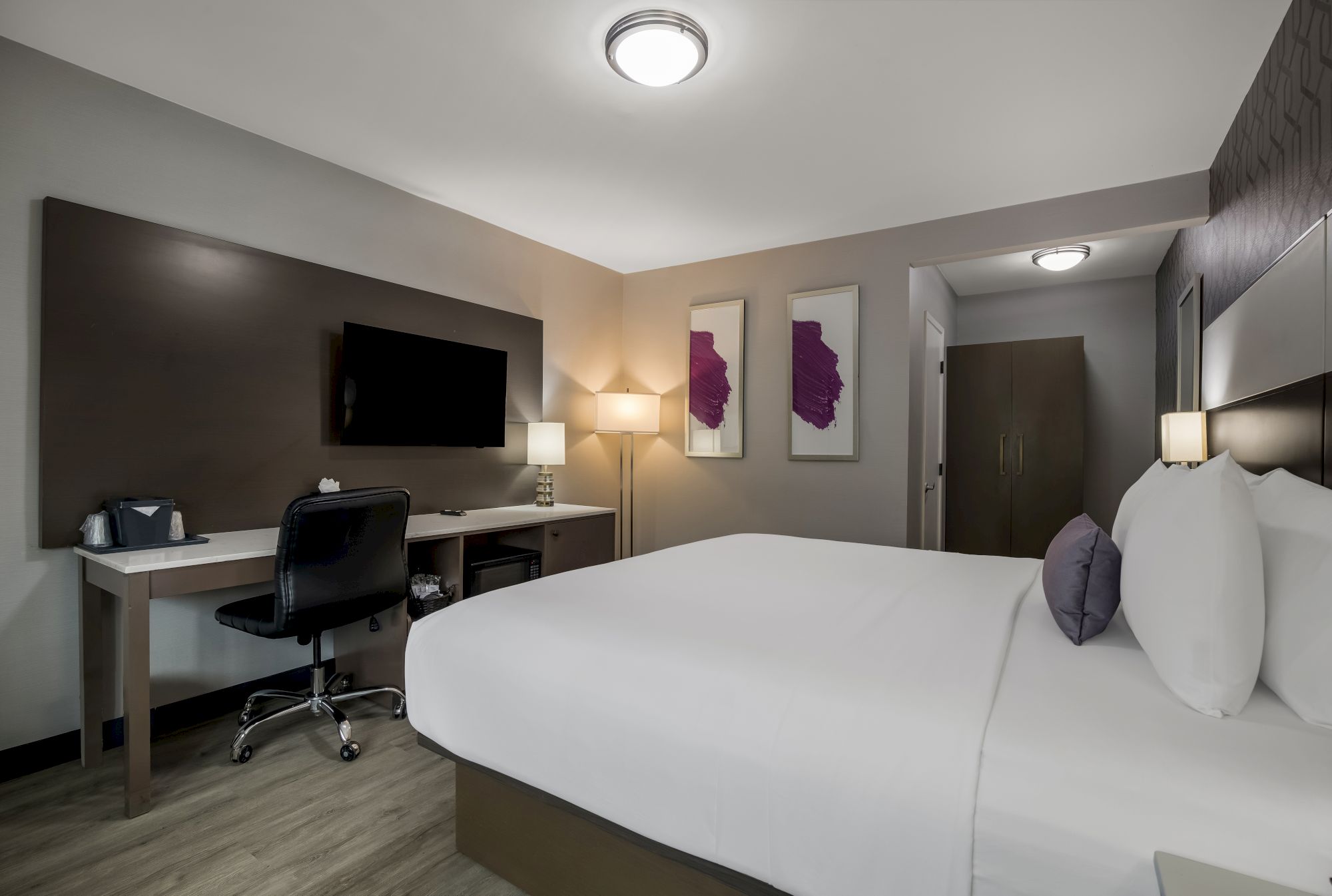 A modern hotel room with a large bed, TV, desk, chair, lamp, and two purple art pieces on the wall. The decor is minimalist and sleek.