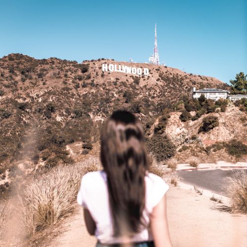 A person with long hair is walking on a dirt path towards the Hollywood Sign, with hills and a clear sky in the background.