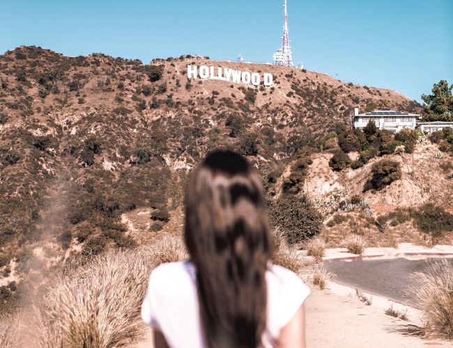 A person with long hair is walking on a dirt path towards the Hollywood Sign, with hills and a clear sky in the background.