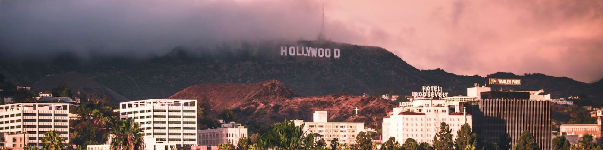 This image shows the Hollywood Sign in Los Angeles, California, partially obscured by clouds, with city buildings and palm trees in the foreground.