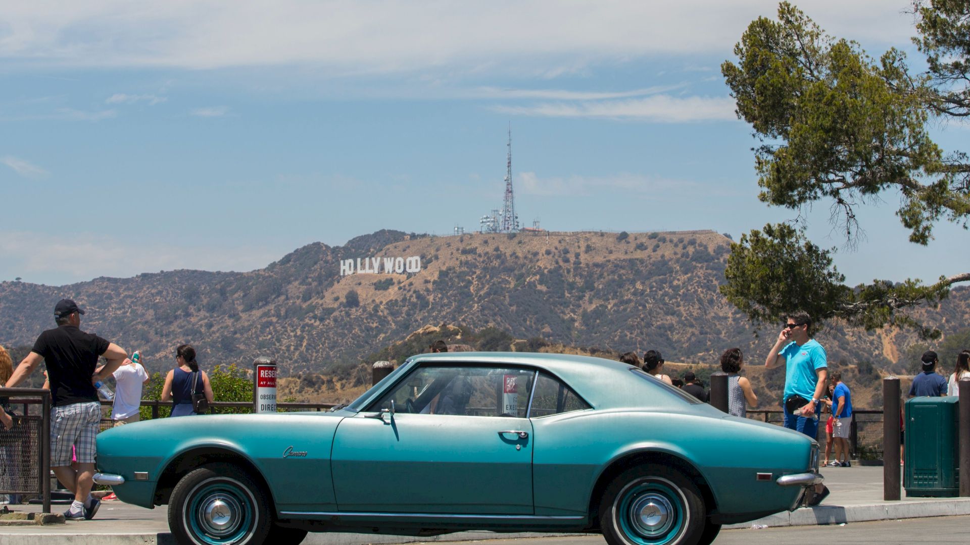 A classic blue car is parked with people nearby, and the iconic "HOLLYWOOD" sign is visible in the background on the hills.