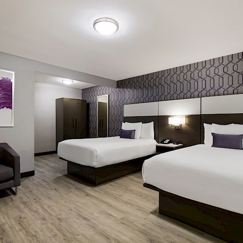 A modern hotel room with two beds, purple cushions, patterned walls, framed artwork, a chair, wardrobe, and wooden flooring, all well-lit.