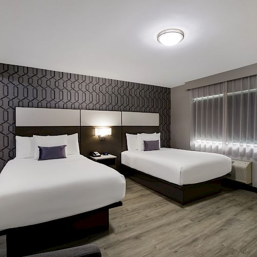 The image shows a modern hotel room with two neatly made beds, geometric wallpaper, a nightstand, a lamp, and a window with closed blinds.