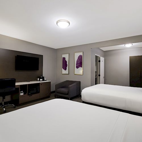 The image shows a hotel room with two beds, a desk with a chair, a TV, an armchair, and wall art, lit by ceiling and desk lamps.