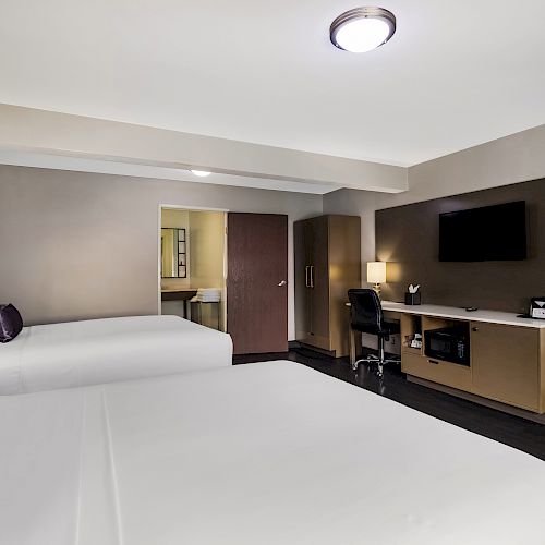 A modern hotel room with two queen beds, a desk, wall-mounted TV, mirror, and a small fridge. There's a door opening to a bathroom.