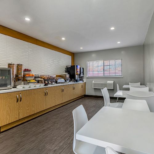 A small dining area with white tables and chairs; a countertop with various breakfast items and beverage machines along one wall.