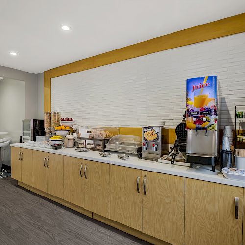 A modern breakfast buffet with a juice dispenser, coffee machine, bread, cereal, and seating area with bar stools.