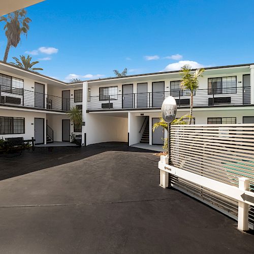 This image shows the courtyard of a two-story motel with outdoor staircases, balcony walkways, and a fenced area, possibly a pool, under a clear blue sky.