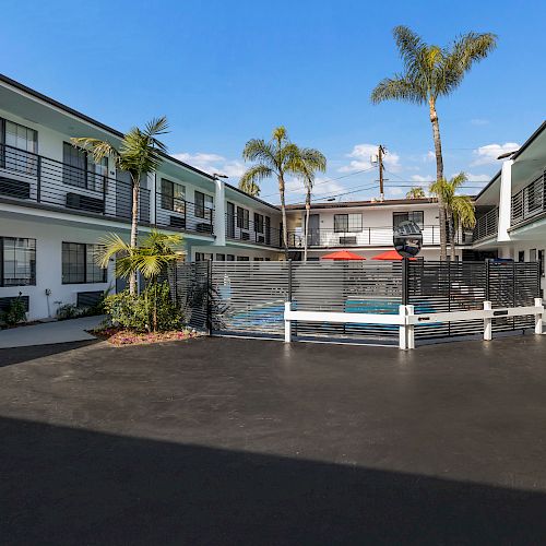 A courtyard enclosed by two-story buildings features palm trees, a fenced swimming pool, and a lounging area in the center.