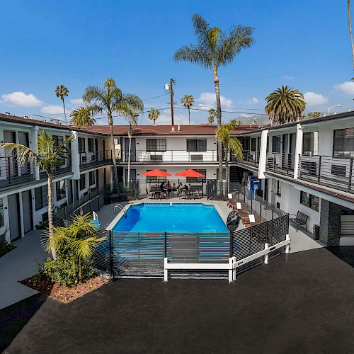 An apartment complex surrounds a central swimming pool, with balconies and palm trees visible in the background under a clear blue sky.