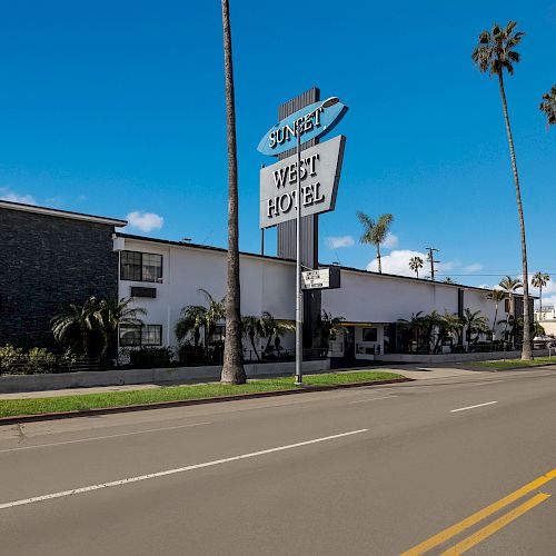 Image of a street with palm trees, a modern two-story building, and a sign reading "Sunset West Hotel." Clear blue sky in the background.