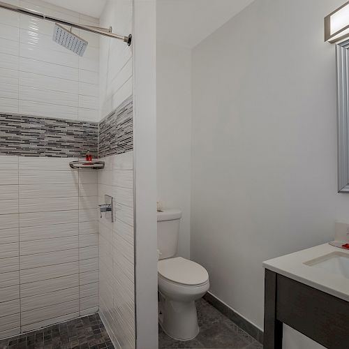 The image shows a modern bathroom with a walk-in shower, a toilet, and a sink with a mirror above it. The design is contemporary with neutral tones.