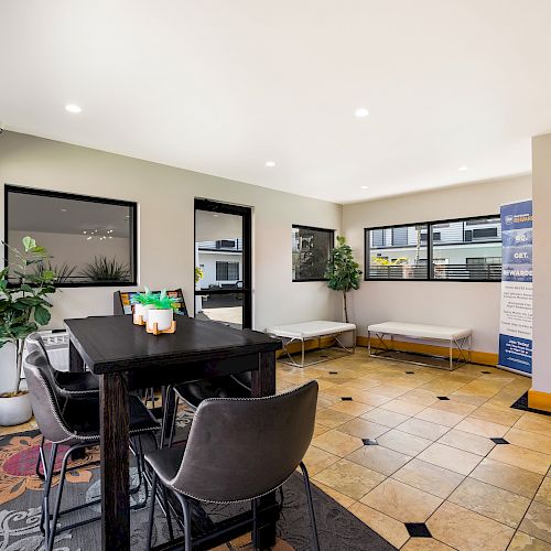 A modern interior space features a dining table with chairs, potted plants, decorative candles, and a large window. The setting is bright and inviting.