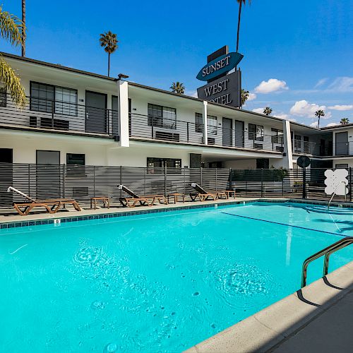 The image shows an outdoor swimming pool surrounded by a modern two-story hotel building with palm trees and lounge chairs under a clear blue sky.