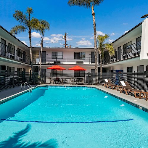 A courtyard pool with lounge chairs, palm trees, and red umbrellas surrounded by a two-story building with balconies, under a clear blue sky.