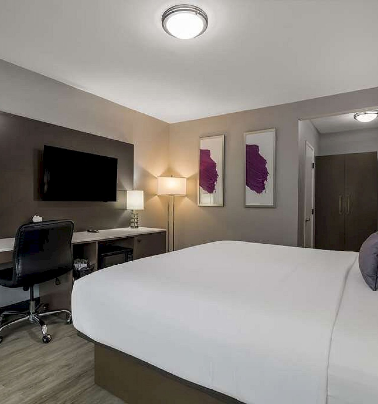 A modern hotel room with a large bed, desk with chair, wall-mounted TV, artwork, and ambient lighting. The room has a minimalist design.