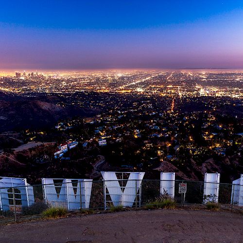 A view of a city at dusk from behind a large, white landmark sign with glowing lights sprawling in the distance.