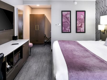 A modern hotel room with a king-sized bed, purple accents, a large TV, a desk, and framed art on the wall.