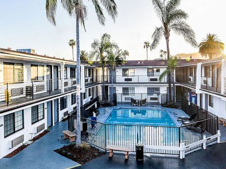 A courtyard of a two-story hotel or apartment complex, featuring a central swimming pool, surrounded by palm trees, fencing, and some outdoor furniture.