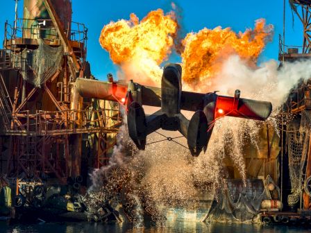 A hydroplane is splashing down on water amid an industrial setting with an explosion in the background, creating a dramatic scene.