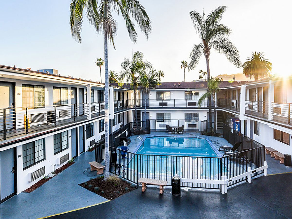 The image shows a two-story motel or apartment complex with a fenced-in swimming pool in the center, surrounded by benches and palm trees.